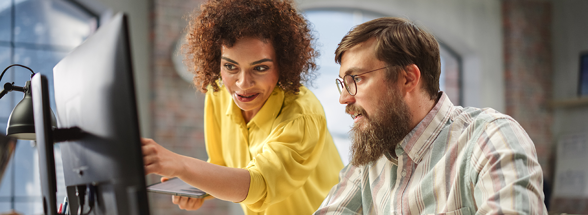 A woman helps her male colleague in an office, showing him something on his computer.