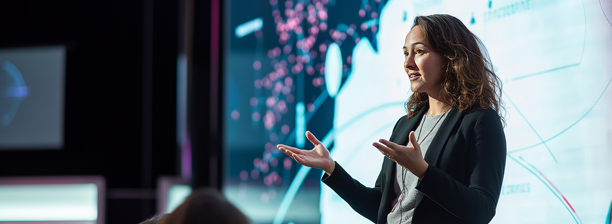 A woman speaker on stage speaking in front of an audience and a large screen.